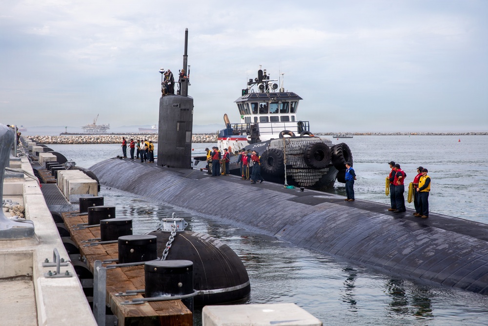 A large military submarine is docking at a naval pier, assisted by a tugboat bearing large rubber fender tires. Numerous crew members in uniform and safety vests are stationed along the submarine's hull and on the dock. The scene appears to be at a U.S. Navy installation, likely on the West Coast based on the breakwater and harbor infrastructure visible in the background.