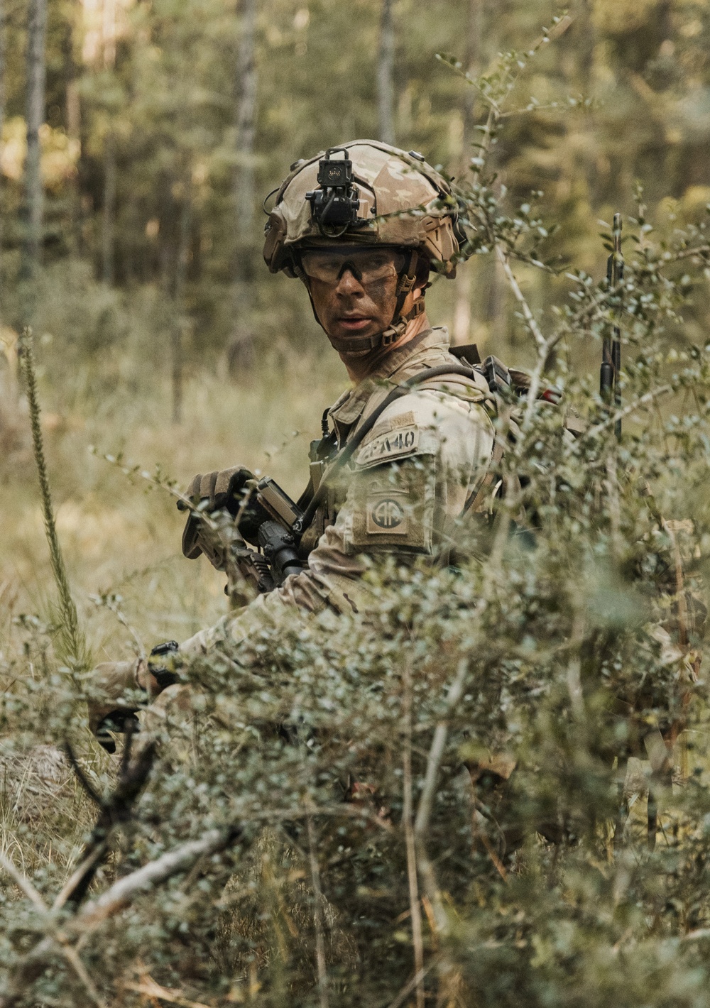 A U.S. Army soldier in full combat gear is positioned among dense woodland vegetation, appearing to be on patrol or in a tactical field exercise. The soldier wears MultiCam camouflage with what appears to be an 82nd Airborne Division patch on the left shoulder, and has face camouflage paint applied. The setting is a forested environment with low scrub brush providing natural concealment.