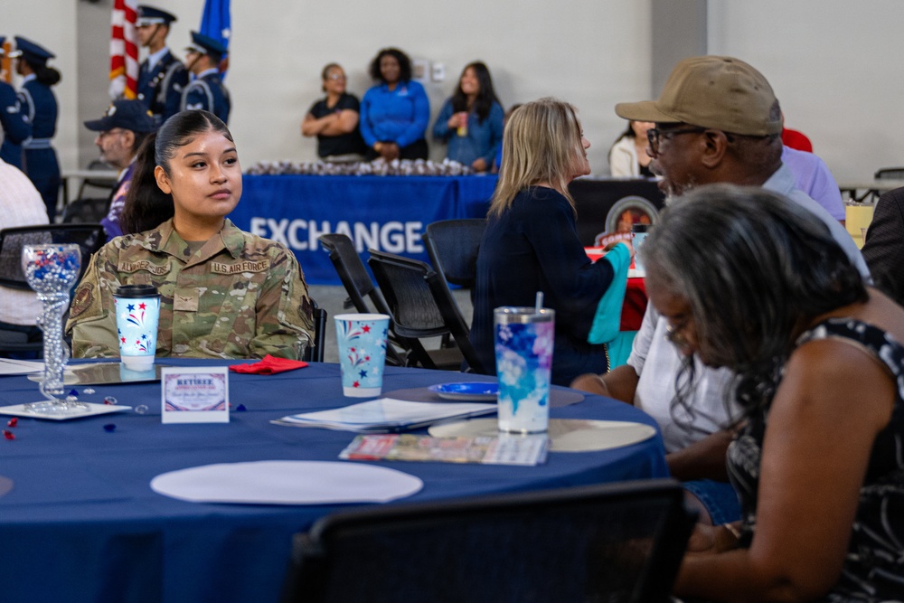A U.S. Air Force service member in OCP (Operational Camouflage Pattern) uniform is seated at a blue-draped table during what appears to be a military appreciation or retiree luncheon event. The setting includes an 'EXCHANGE' banner in the background and a color guard formation visible in the rear. The event appears to be a formal military community gathering with multiple attendees seated at decorated tables.
