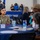 A U.S. Air Force service member in OCP (Operational Camouflage Pattern) uniform is seated at a blue-draped table during what appears to be a military appreciation or retiree luncheon event. The setting includes an 'EXCHANGE' banner in the background and a color guard formation visible in the rear. The event appears to be a formal military community gathering with multiple attendees seated at decorated tables.