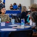 A U.S. Air Force service member in OCP (Operational Camouflage Pattern) uniform is seated at a blue-draped table during what appears to be a military appreciation or retiree luncheon event. The setting includes an 'EXCHANGE' banner in the background and a color guard formation visible in the rear. The event appears to be a formal military community gathering with multiple attendees seated at decorated tables.