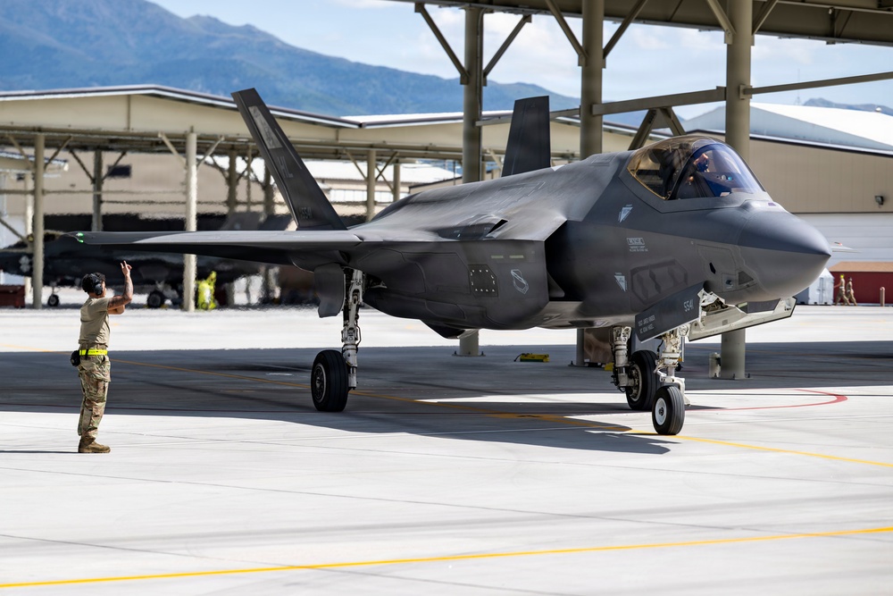 A military F-35 Lightning II stealth fighter jet is taxiing on an airfield tarmac while a ground crew member in military uniform directs the aircraft using hand signals. The scene takes place at what appears to be a military air base with large hangars and mountainous terrain visible in the background. The aircraft is in a dark gray stealth finish with landing gear deployed.