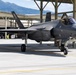 A military F-35 Lightning II stealth fighter jet is taxiing on an airfield tarmac while a ground crew member in military uniform directs the aircraft using hand signals. The scene takes place at what appears to be a military air base with large hangars and mountainous terrain visible in the background. The aircraft is in a dark gray stealth finish with landing gear deployed.