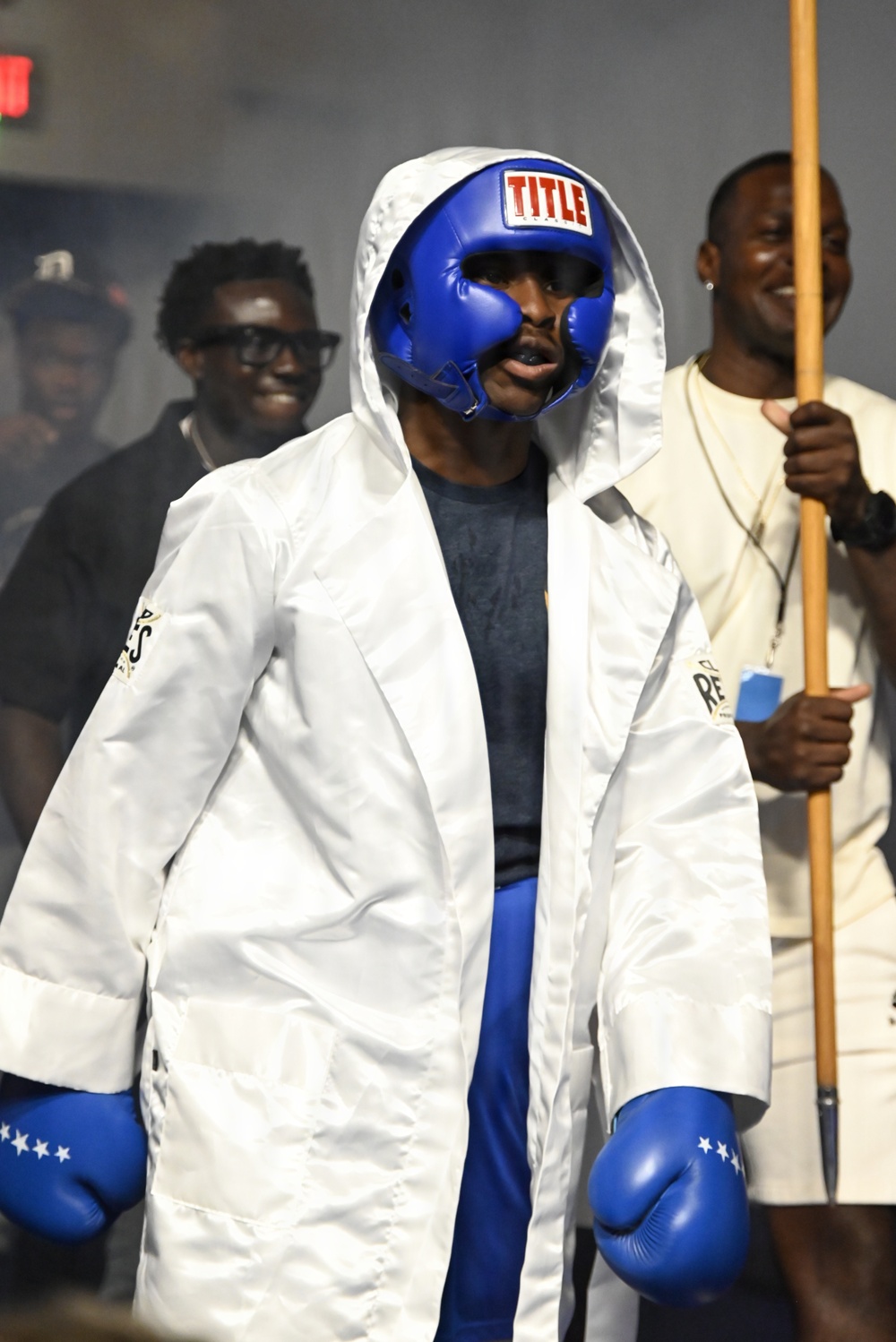 A boxer is shown making a ring entrance wearing a blue 'TITLE' brand headguard, blue boxing gloves with white stars, and a white hooded robe. The fighter appears focused and ready for competition, accompanied by corner team members in the background. The setting appears to be an indoor boxing event with atmospheric smoke effects.