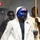 A boxer is shown making a ring entrance wearing a blue 'TITLE' brand headguard, blue boxing gloves with white stars, and a white hooded robe. The fighter appears focused and ready for competition, accompanied by corner team members in the background. The setting appears to be an indoor boxing event with atmospheric smoke effects.