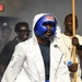 A boxer is shown making a ring entrance wearing a blue 'TITLE' brand headguard, blue boxing gloves with white stars, and a white hooded robe. The fighter appears focused and ready for competition, accompanied by corner team members in the background. The setting appears to be an indoor boxing event with atmospheric smoke effects.