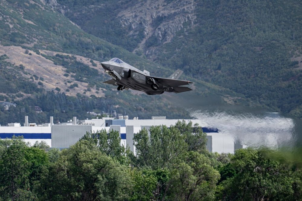 A military stealth fighter jet, identifiable as an F-35 Lightning II, is captured in low-altitude flight with landing gear deployed, suggesting a takeoff or landing phase. The aircraft is photographed against a backdrop of tree-covered mountains, industrial or military buildings, and what appears to be a large body of water. The image appears to be taken near a military air base situated in a mountainous valley.