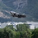 A military stealth fighter jet, identifiable as an F-35 Lightning II, is captured in low-altitude flight with landing gear deployed, suggesting a takeoff or landing phase. The aircraft is photographed against a backdrop of tree-covered mountains, industrial or military buildings, and what appears to be a large body of water. The image appears to be taken near a military air base situated in a mountainous valley.