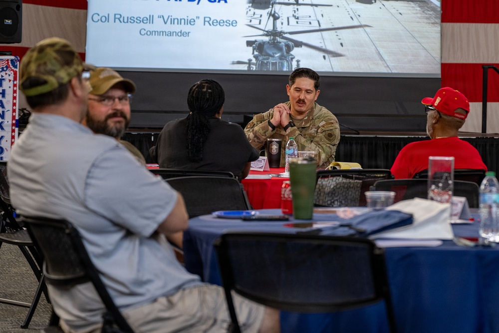 A military event or briefing is taking place in a decorated venue with American flag bunting. A uniformed service member in OCP (Operational Camouflage Pattern) uniform is seated at a table engaging with civilians. A presentation slide in the background identifies 'Col Russell "Vinnie" Reese, Commander' alongside an image of a military helicopter.