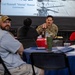 A military event or briefing is taking place in a decorated venue with American flag bunting. A uniformed service member in OCP (Operational Camouflage Pattern) uniform is seated at a table engaging with civilians. A presentation slide in the background identifies 'Col Russell "Vinnie" Reese, Commander' alongside an image of a military helicopter.