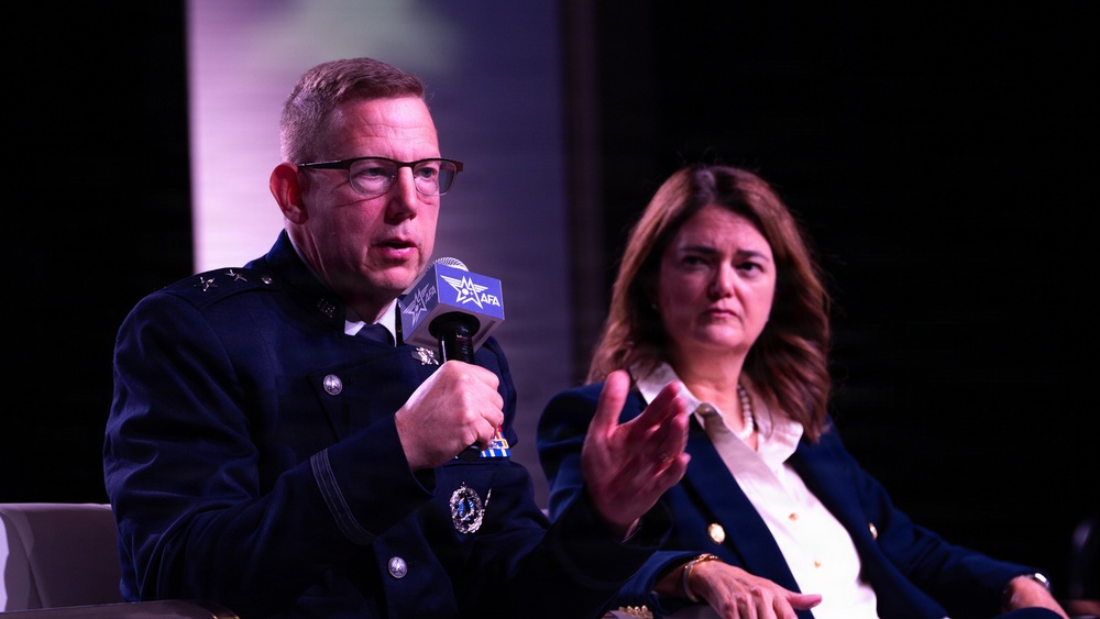 A military officer in dress uniform is speaking into an AFA (Air Force Association) branded microphone at what appears to be a panel discussion or conference. A woman in a navy blazer sits beside him, listening attentively. The setting appears to be a formal event stage with dark background lighting.