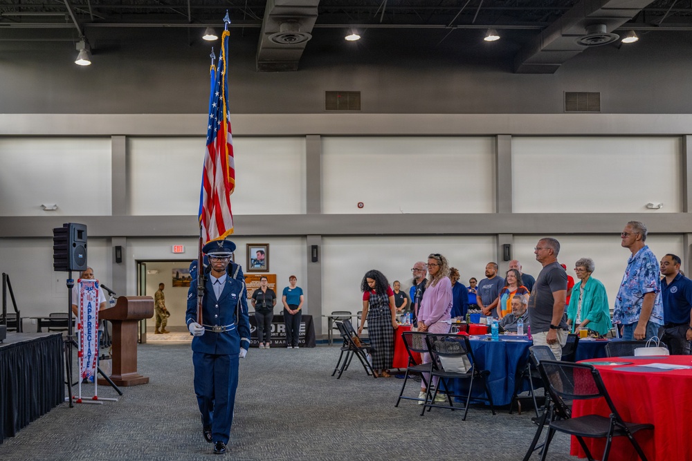 A military color guard ceremony is taking place inside a large indoor venue, likely a convention center or military facility. A uniformed Air Force service member is carrying the United States flag while attendees stand respectfully at attention. The event appears to be a formal military appreciation or recognition ceremony.