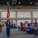 A military color guard ceremony is taking place inside a large indoor venue, likely a convention center or military facility. A uniformed Air Force service member is carrying the United States flag while attendees stand respectfully at attention. The event appears to be a formal military appreciation or recognition ceremony.