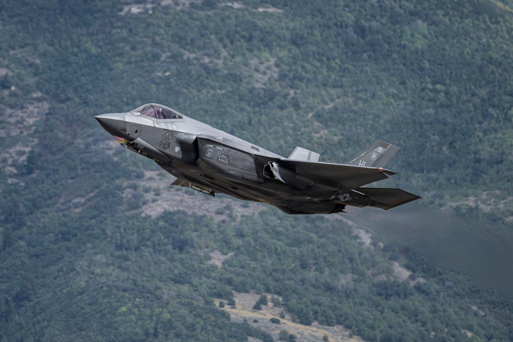 A single-seat stealth fighter jet, identifiable as an F-35 Lightning II, is captured in low-altitude flight against a backdrop of densely forested mountainous terrain. The aircraft appears to be in a climbing or banking maneuver, with landing gear retracted and weapons visible on underwing hardpoints. The image is sharp and well-composed, suggesting a professional aviation or military photography context.