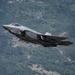 A single-seat stealth fighter jet, identifiable as an F-35 Lightning II, is captured in low-altitude flight against a backdrop of densely forested mountainous terrain. The aircraft appears to be in a climbing or banking maneuver, with landing gear retracted and weapons visible on underwing hardpoints. The image is sharp and well-composed, suggesting a professional aviation or military photography context.