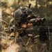 A soldier in full combat gear is lying prone in a wooded area, aiming a rifle equipped with an optical scope. The individual is wearing camouflage uniform, a tactical helmet with mounted devices including what appears to be a small action camera and night vision mount. The setting appears to be a forest training or operational environment with natural ground cover.