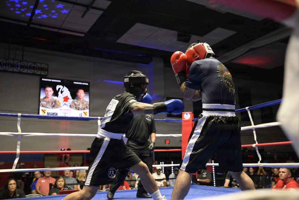 Two boxers are actively competing in a boxing ring during what appears to be a military-themed boxing event called 'Phantom Warriors.' The fighter on the left wearing black shorts and blue gloves is throwing a punch at the opponent on the right who is wearing red gloves and a dark blue outfit. A crowd of spectators is visible in the background along with a display screen showing fighter profiles.