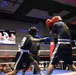 Two boxers are actively competing in a boxing ring during what appears to be a military-themed boxing event called 'Phantom Warriors.' The fighter on the left wearing black shorts and blue gloves is throwing a punch at the opponent on the right who is wearing red gloves and a dark blue outfit. A crowd of spectators is visible in the background along with a display screen showing fighter profiles.