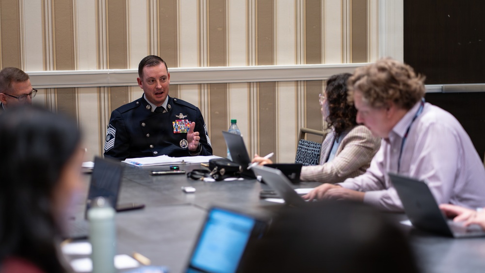 A military officer in dress uniform, identifiable by rank insignia consistent with a Chief Master Sergeant, is speaking at a conference or meeting table surrounded by civilians with laptops. The setting appears to be a hotel or conference room with striped wallpaper. The meeting appears to be a formal briefing or panel discussion involving both military and civilian participants.