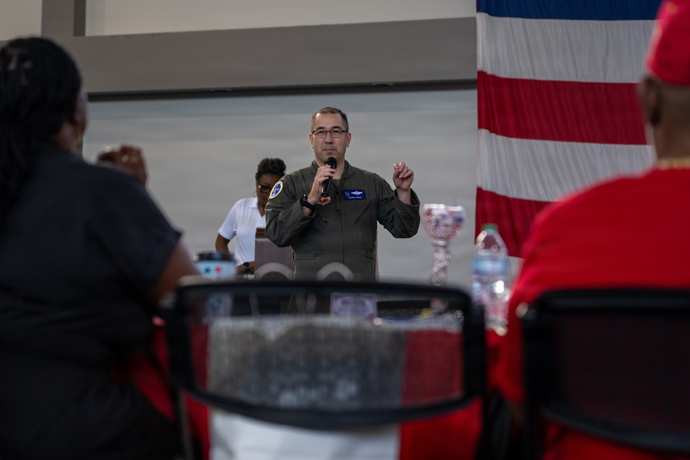 A military officer in an olive green flight suit is speaking into a microphone while addressing an audience. He appears to be giving a presentation or speech at what looks like an official military event or ceremony. An American flag is visible in the background to the right.