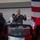A military officer in an olive green flight suit is speaking into a microphone while addressing an audience. He appears to be giving a presentation or speech at what looks like an official military event or ceremony. An American flag is visible in the background to the right.