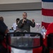 A military officer in an olive green flight suit is speaking into a microphone while addressing an audience. He appears to be giving a presentation or speech at what looks like an official military event or ceremony. An American flag is visible in the background to the right.