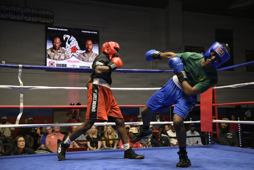 Two boxers are competing in a boxing ring during what appears to be a military boxing event called 'Phantom Warriors vs Phantom Lethal.' One boxer wearing red gear and 'Title Boxing' shorts is exchanging punches with an opponent in blue gear and a green shirt. A crowd of spectators watches from ringside, and a scoreboard/display screen in the background shows fighter profiles labeled 'Webb' and 'Jackson' in the Middleweight category.