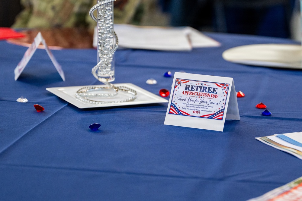 A decorated banquet table set up for a 'Retiree Appreciation Day' event, featuring patriotic red, white, and blue decorations. The table displays a tent card thanking retirees for their service, along with decorative gemstones and a silver centerpiece. The setting suggests a formal military or government retiree recognition ceremony.