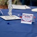 A decorated banquet table set up for a 'Retiree Appreciation Day' event, featuring patriotic red, white, and blue decorations. The table displays a tent card thanking retirees for their service, along with decorative gemstones and a silver centerpiece. The setting suggests a formal military or government retiree recognition ceremony.