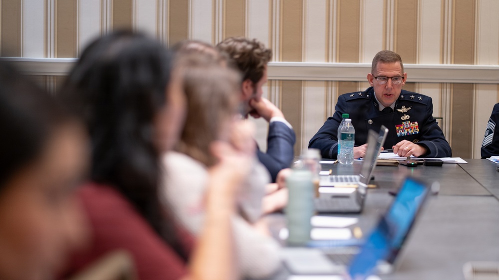 A military officer in a dark blue dress uniform with two stars and multiple medals is speaking at a conference table to a group of civilians. The setting appears to be a formal briefing or meeting room, with attendees seated along the table with laptops and documents. The foreground figures are intentionally blurred, keeping focus on the uniformed speaker.