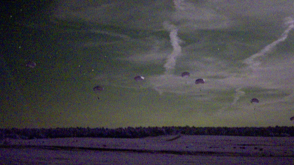 A low-light or night-vision style photograph showing multiple parachutes deployed in the sky over a flat, open landscape. The image has a distinctive green-purple tonal quality consistent with either night photography or a color-shifted film/sensor. Several parachutes are visible at varying altitudes, suggesting an airborne drop operation.