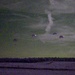 A low-light or night-vision style photograph showing multiple parachutes deployed in the sky over a flat, open landscape. The image has a distinctive green-purple tonal quality consistent with either night photography or a color-shifted film/sensor. Several parachutes are visible at varying altitudes, suggesting an airborne drop operation.