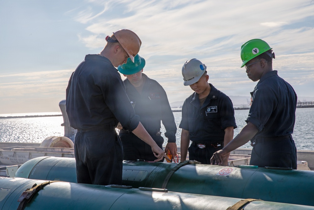 Four military personnel in dark navy coveralls are working together on what appears to be a torpedo or similar cylindrical ordnance on the deck of a naval vessel. The scene takes place outdoors near open water, with the personnel wearing safety equipment including hard hats and protective eyewear. The team appears to be conducting maintenance, inspection, or assembly procedures on the weapon system.