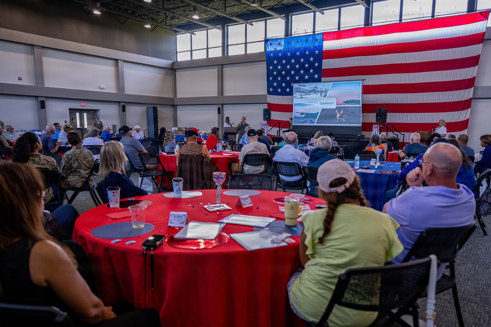 A large indoor gathering is taking place in a modern hall decorated with an oversized American flag as a backdrop. Attendees are seated at round tables with red tablecloths, watching a presentation displayed on a projection screen. The event appears to be a military or patriotic community briefing or luncheon.