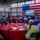 A large indoor gathering is taking place in a modern hall decorated with an oversized American flag as a backdrop. Attendees are seated at round tables with red tablecloths, watching a presentation displayed on a projection screen. The event appears to be a military or patriotic community briefing or luncheon.