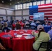 A large indoor gathering is taking place in a modern hall decorated with an oversized American flag as a backdrop. Attendees are seated at round tables with red tablecloths, watching a presentation displayed on a projection screen. The event appears to be a military or patriotic community briefing or luncheon.