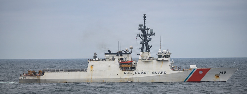 A U.S. Coast Guard cutter with hull number 755 is underway on open ocean waters under an overcast sky. The large white vessel displays the distinctive Coast Guard diagonal stripe in red and blue on its bow. Light smoke is visible from the vessel's exhaust, indicating active propulsion.