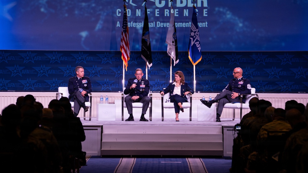 A panel discussion is taking place on a stage at an AFA (Air Force Association) Cyber Conference, described as a Professional Development Event. Four panelists — three in military uniforms and one in civilian attire — are seated in white chairs on a raised white platform. An audience is visible in the foreground watching the presentation.