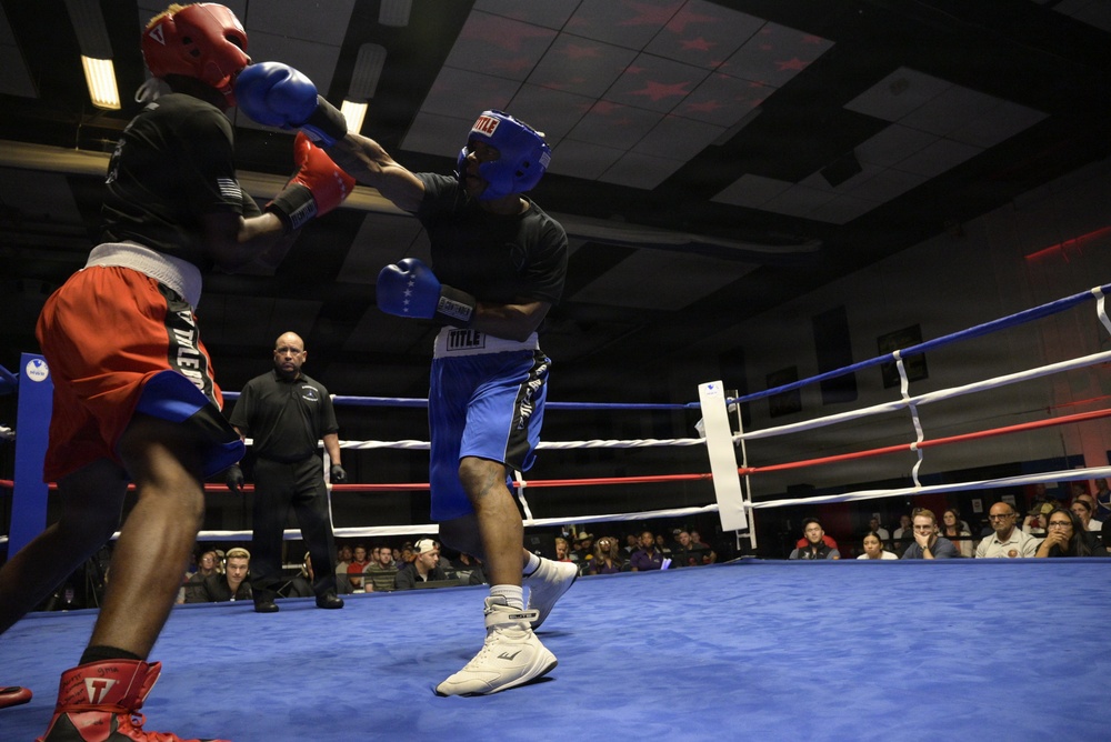 Two boxers are engaged in an active match inside a boxing ring with blue canvas flooring. One boxer wears red headgear and red/orange shorts, while the other wears blue headgear and blue shorts, both using TITLE brand equipment. A referee in black monitors the bout as a large crowd of spectators watches from the background.