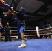Two boxers are engaged in an active match inside a boxing ring with blue canvas flooring. One boxer wears red headgear and red/orange shorts, while the other wears blue headgear and blue shorts, both using TITLE brand equipment. A referee in black monitors the bout as a large crowd of spectators watches from the background.