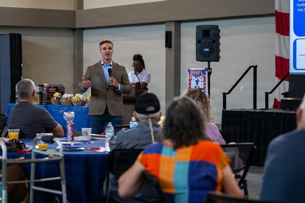 A man in a tan blazer and jeans is speaking into a microphone at what appears to be a community or veterans-related event. Attendees are seated at round tables with blue tablecloths, listening to the presentation. The setting includes patriotic decorations, a PA speaker system, and a projection screen in the background.