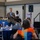 A man in a tan blazer and jeans is speaking into a microphone at what appears to be a community or veterans-related event. Attendees are seated at round tables with blue tablecloths, listening to the presentation. The setting includes patriotic decorations, a PA speaker system, and a projection screen in the background.