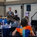 A man in a tan blazer and jeans is speaking into a microphone at what appears to be a community or veterans-related event. Attendees are seated at round tables with blue tablecloths, listening to the presentation. The setting includes patriotic decorations, a PA speaker system, and a projection screen in the background.