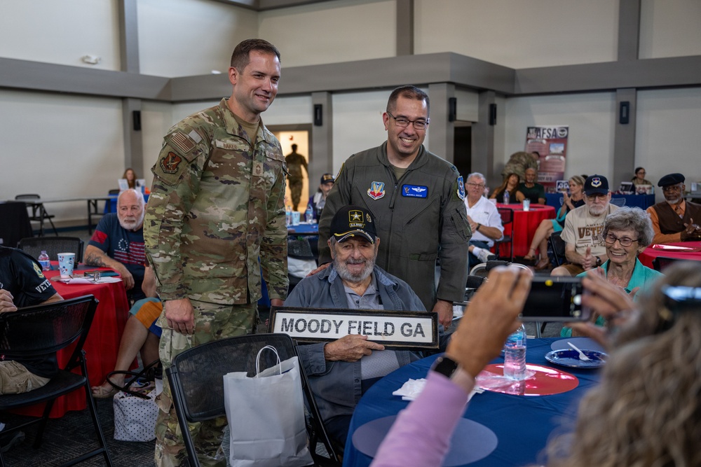 A veteran seated in a chair holds a sign reading 'MOODY FIELD GA' while two active-duty military personnel stand behind him posing for a photo. The event appears to be a veterans appreciation gathering or ceremony held in a banquet/community hall setting. Multiple other veterans and guests are seated at decorated tables in the background.