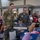 A veteran seated in a chair holds a sign reading 'MOODY FIELD GA' while two active-duty military personnel stand behind him posing for a photo. The event appears to be a veterans appreciation gathering or ceremony held in a banquet/community hall setting. Multiple other veterans and guests are seated at decorated tables in the background.