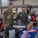 A veteran seated in a chair holds a sign reading 'MOODY FIELD GA' while two active-duty military personnel stand behind him posing for a photo. The event appears to be a veterans appreciation gathering or ceremony held in a banquet/community hall setting. Multiple other veterans and guests are seated at decorated tables in the background.