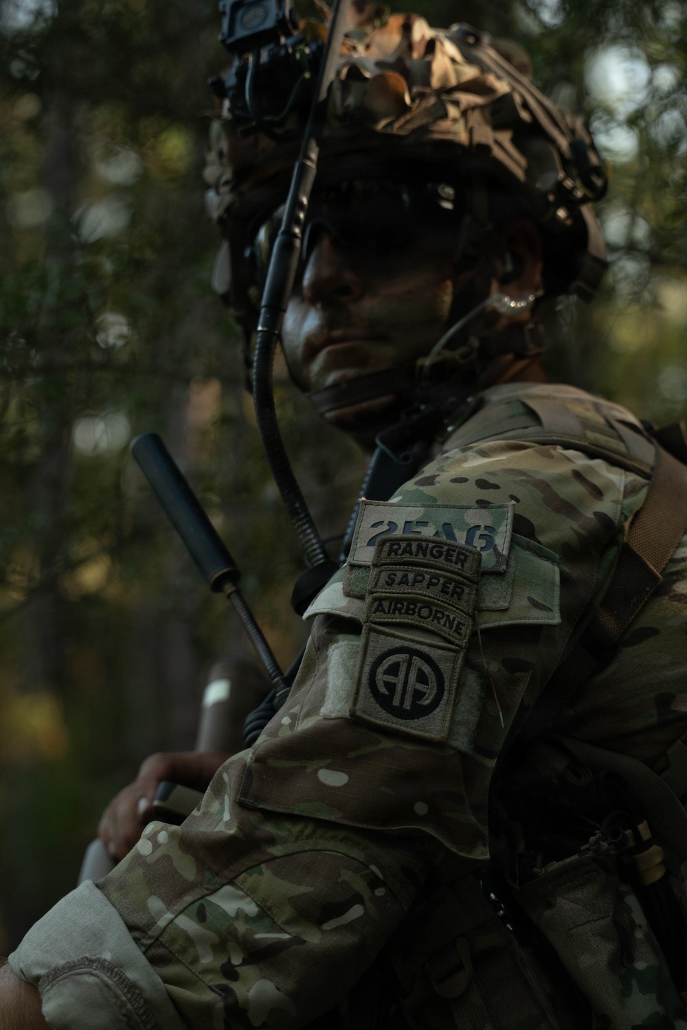A U.S. Army soldier in full combat gear is photographed in a wooded environment. The soldier wears MultiCam camouflage and displays multiple qualification tabs and unit patches on the left shoulder. The image has a dramatic, low-light aesthetic with bokeh background.