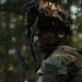 A U.S. Army soldier in full combat gear is photographed in a wooded environment. The soldier wears MultiCam camouflage and displays multiple qualification tabs and unit patches on the left shoulder. The image has a dramatic, low-light aesthetic with bokeh background.