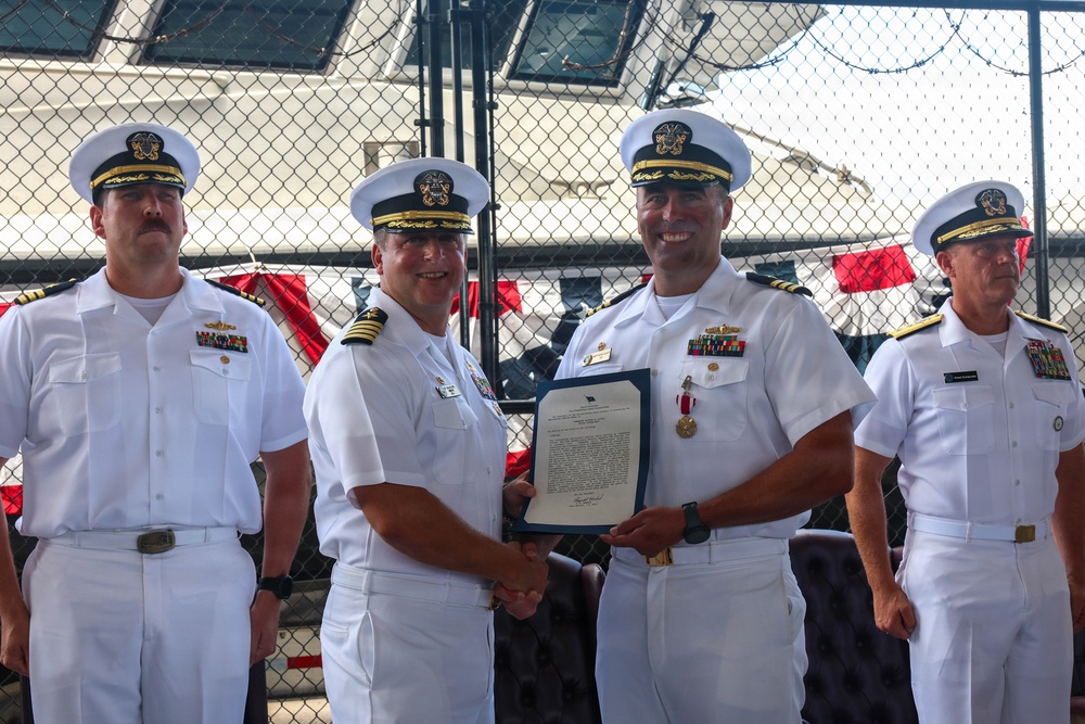 Four U.S. Navy officers in white dress uniforms are participating in a formal military ceremony, likely a change of command or award presentation. Two central figures are shaking hands while jointly holding an official document in a blue folder, with a medal visible on one officer's chest. The event appears to take place on or near a naval vessel, with chain-link fencing and ship structures visible in the background.