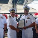 Four U.S. Navy officers in white dress uniforms are participating in a formal military ceremony, likely a change of command or award presentation. Two central figures are shaking hands while jointly holding an official document in a blue folder, with a medal visible on one officer's chest. The event appears to take place on or near a naval vessel, with chain-link fencing and ship structures visible in the background.